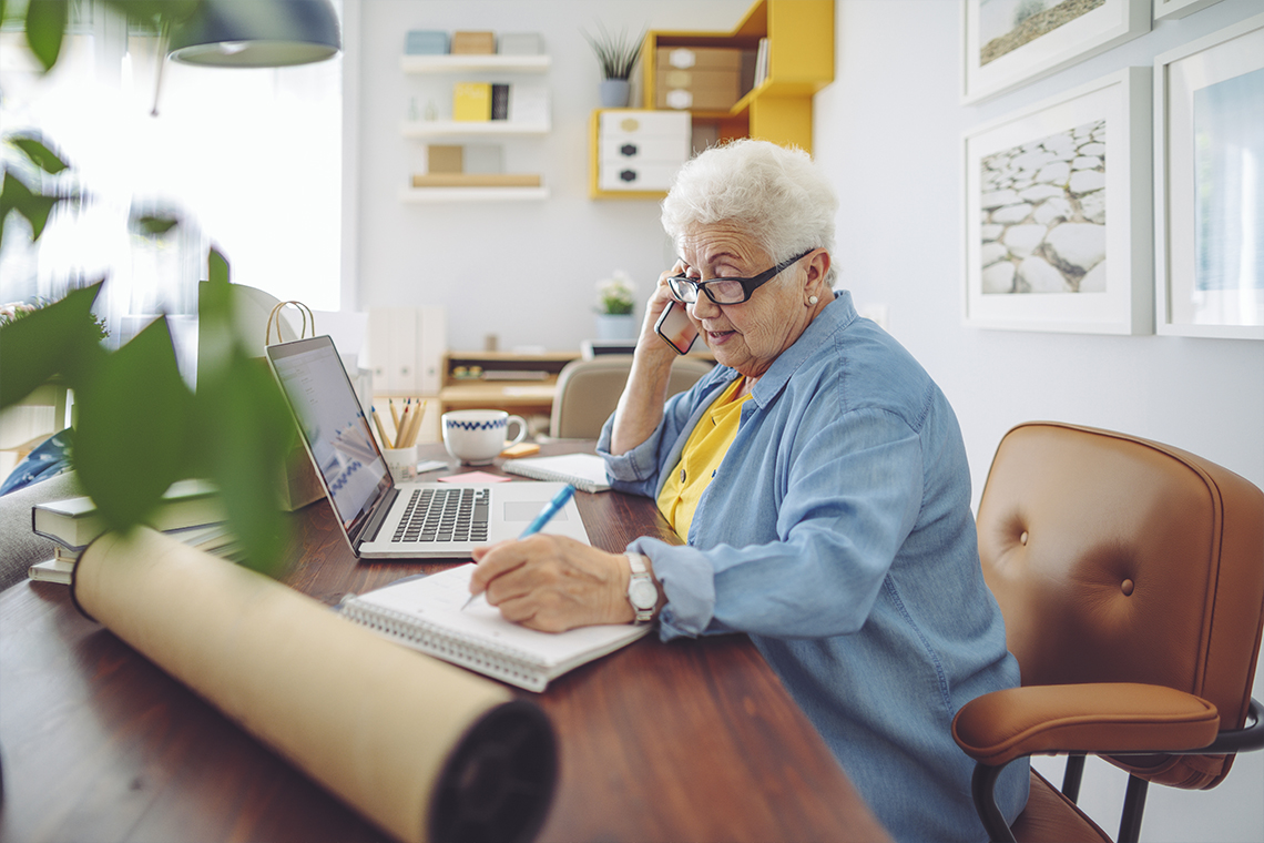 Pensionär med vitt hår och glasögon som antecknar i ett block bredvid sin laptop.